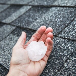 person holding large hail stone in hand in front of roof shingles
